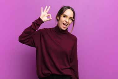 Genç, güzel bir kadın kendini başarılı ve tatmin olmuş hissediyor. Ağzı açık gülümsüyor ve elini mor duvara yaslayarak işaret diliyle işaret veriyor.