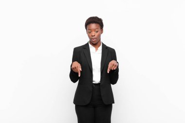 Genç, güzel, siyah bir kadın, ağzı açık, iki eliyle aşağıya bakıyor, şaşkın, şaşkın ve şaşırmış görünüyor beyaz duvara karşı