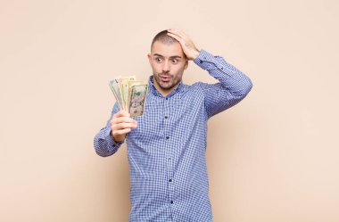 Genç yakışıklı adam mutlu, şaşkın ve şaşırmış görünüyor, gülümsüyor ve dolar banknotlarıyla inanılmaz ve inanılmaz iyi haberler veriyor.