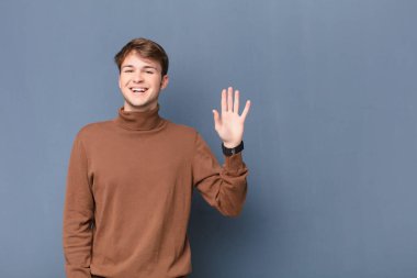 Genç sarışın adam mutlu ve mutlu bir şekilde gülümsüyor, el sallıyor, seni selamlıyor ve selamlıyor, ya da düz duvara yaslanmış bir şekilde veda ediyor.