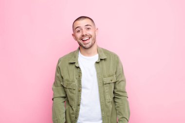 Genç, yakışıklı, büyük, dost canlısı, kaygısız bir gülümsemesi olan, pozitif görünen, rahat ve mutlu görünen, düz duvara karşı ürperen bir adam.