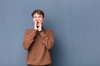 Genç sarışın adam mutlu, heyecanlı ve pozitif hissediyor, elleri ağızında bağırıyor, duvara yaslanmış bir şekilde sesleniyor.