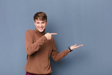 Genç sarışın adam neşeyle gülümsüyor ve avuç içinin kenarındaki fotokopi alanını işaret ediyor. Duvara yaslanmış bir nesneyi gösteriyor ya da reklamını yapıyor.