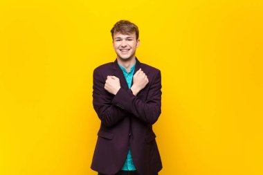 Sarışın genç adam sevinçle gülümsüyor ve kollarını kavuşturup yumruklarını sıkıyor, mutlu ve pozitif bir şekilde yassı duvara yaslanıyor.