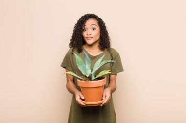 Genç siyah kadın şaşkın ve şaşkın hissediyor, şüpheci, farklı seçenekleri tartıyor ya da seçiyor elinde kaktüs tutan komik bir ifadeyle.
