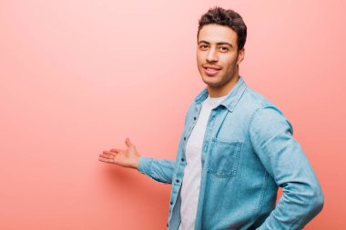 Genç Arap adam kendini mutlu ve neşeli hissediyor, gülümsüyor ve seni hoş karşılıyor, pembe duvara karşı dostça bir jestle içeri davet ediyor.