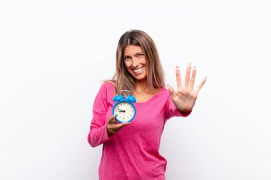young pretty woman smiling and looking friendly, showing number five or fifth with hand forward, counting down holding an alarm clock. clipart