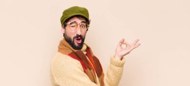Genç havalı sakallı adam şok olmuş, şaşırmış ve şaşırmış hissediyor. Onaylıyor. İki eliyle işaret veriyor.