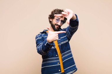 Genç çılgın havalı adam mutlu, arkadaş canlısı ve pozitif hissediyor, gülümsüyor ve elleri düz duvara dayalı bir portre veya fotoğraf çerçevesi yapıyor.