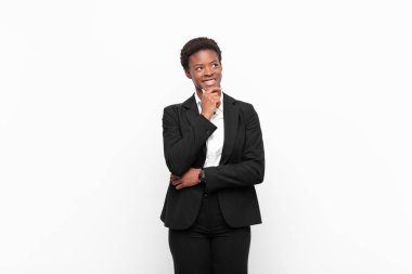 Genç, güzel, siyah kadın gülümseyen mutlu, kendine güveni tam, eli çenesinde, beyaz duvara bakıp merak eden ve yan tarafa bakan.