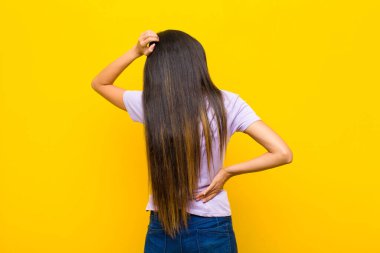 Genç, güzel, Latin bir kadın bilgisiz ve kafası karışık, bir çözüm düşünüyor, eli kalçasında ve diğerinin kafasında, düz duvara bakan dikiz aynası