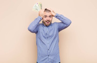 Genç, yakışıklı bir adam stresli, endişeli, endişeli ya da korkmuş, elleri başının üstünde, dolar banknotlarıyla yanlışlıkla panikliyor.
