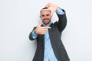 Genç işadamı mutlu, arkadaş canlısı ve pozitif hissediyor, gülümsüyor ve elleri düz duvara dayalı bir portre veya fotoğraf çerçevesi yapıyor.