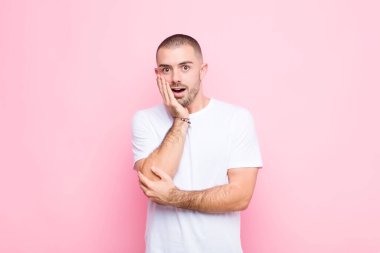 Genç yakışıklı adam şok ve inançsızlık içinde, elleri yanak ve kolları çapraz, sersemlemiş ve düz duvara karşı şaşkın hissediyor