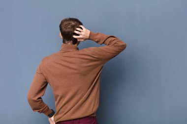 Genç, sarışın bir adam bilgisiz ve şaşkın hissediyor, bir çözüm düşünüyor, eli kalçasında, diğeri kafasında, dikiz aynası düz duvara karşı izole.