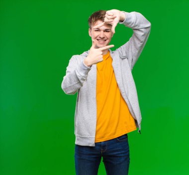 Genç sarışın adam mutlu, arkadaş canlısı ve pozitif hissediyor, gülümsüyor ve elleri kroma tuşuna dayalı bir portre veya fotoğraf çerçevesi yapıyor.