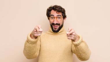 Genç havalı adam mutlu ve kendinden emin, kamerayı iki eliyle gösteriyor ve gülüyor, seni düz duvara karşı seçiyor.