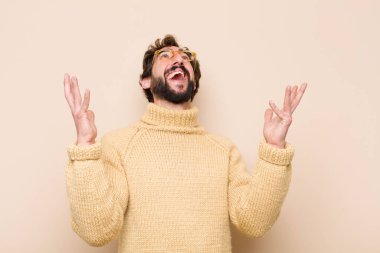 Genç havalı adam kendini mutlu, şaşkın, şanslı ve şaşırmış hissediyor, zaferi kutlarken iki elini de havaya kaldırmış düz duvara karşı.