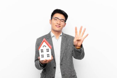 Genç Japon adam gülümsüyor ve arkadaş canlısı görünüyor, dört ya da dört numarayı gösteriyor, bir ev modeliyle geriye doğru sayıyor.