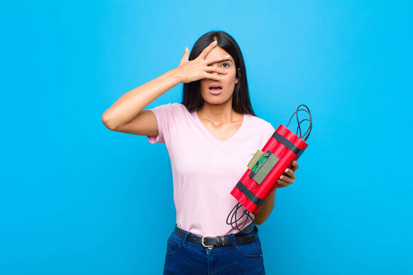 молодая красивая латинская женщина, выглядящая шокированной, напуганной или напуганной, закрывающая лицо рукой и подглядывающая между пальцами к плоской стене