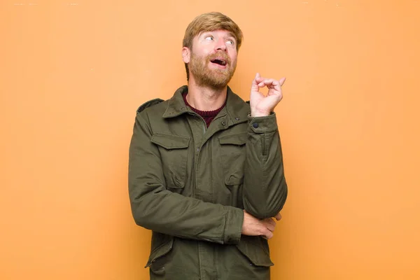 young blonde man smiling happily and looking sideways, wondering, thinking or having an idea against flat wall