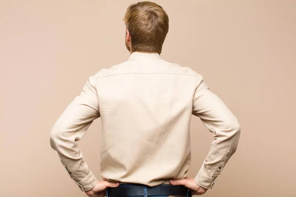 young blonde man feeling confused or full or doubts and questions, wondering, with hands on hips, rear view against flat wall