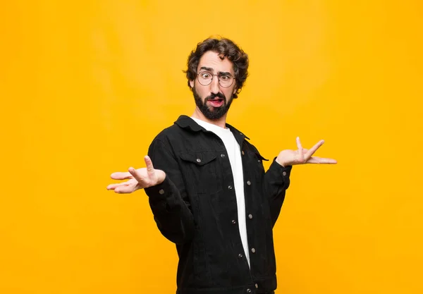 young crazy handsome man shrugging with a dumb, crazy, confused, puzzled expression, feeling annoyed and clueless against orange wall