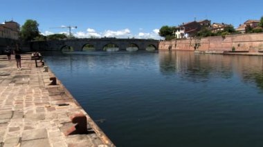 Eski Köprü Tiberius (Ponte di Tiberio), Rimini, İtalya için arka plan ile riverside tarafından insanlar yürümek.