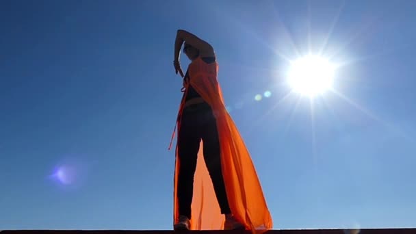 Vacker modell stående i solen lyser på den blå bakgrunden. Vinden blåser och utvecklar hennes mantel. Slow motion. — Stockvideo