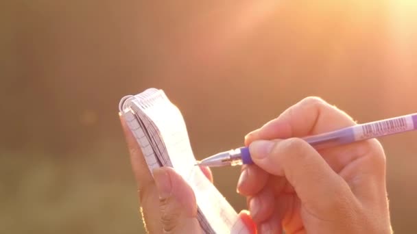 Το κορίτσι του χέρι κάνοντας σημειώσεις σε ένα σημειωματάριο στο ηλιοβασίλεμα σε ακτίνες του ήλιου. — Αρχείο Βίντεο