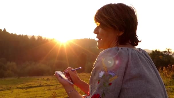 Mulher escrevendo notas em seu bloco de notas ao pôr do sol e sorrindo no final . — Vídeo de Stock