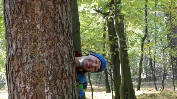 Um jovem espreitando e inclinando a mão por trás de um pinheiro . — Vídeo de Stock