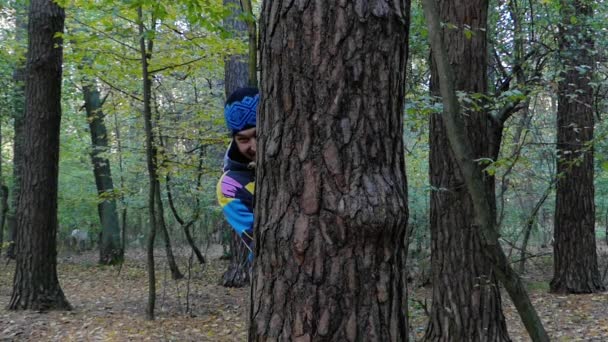 Un jeune homme regardant derrière un arbre et agitant la main dans la forêt d'automne . — Video