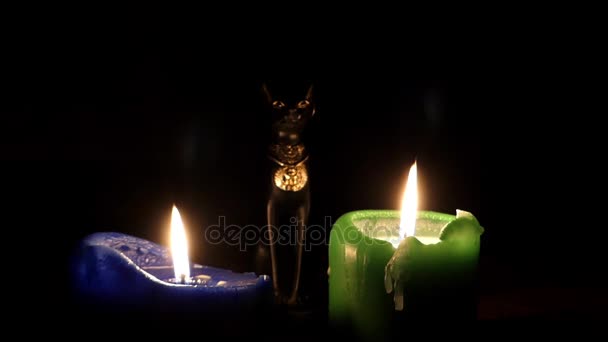 Uma vela azul e uma verde e uma estatueta de gato preto entre elas à noite . — Vídeo de Stock
