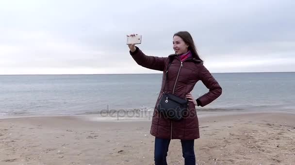 En ung, vakker jente smiler og tar en Selfie mens hun er på Sandy Beach om høsten. – stockvideo