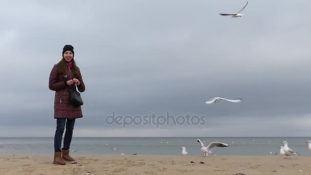 Una joven sonríe y alimenta a las gaviotas en un fondo marino con un clima ventoso en Slo-Mo — Vídeos de Stock