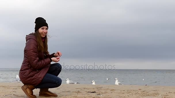 En ung leende flicka sitter och matar fiskmåsar på en sandstrand i höst Slo-Mo — Stockvideo
