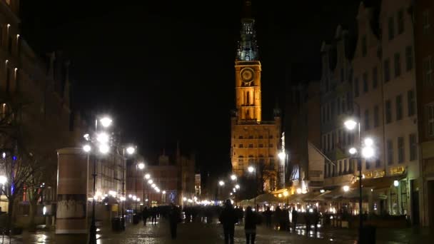 Een Pools stadsplein met een hoge oude toren bij nacht en mensen lopen langs het. — Stockvideo