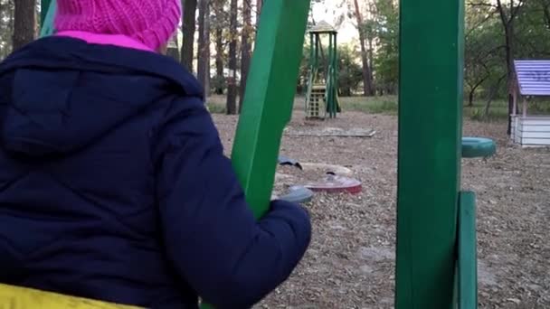 Una mujer en una parka negra y un sombrero de punto rosa se sienta en un columpio de madera en movimiento en un parque infantil del bosque — Vídeo de stock