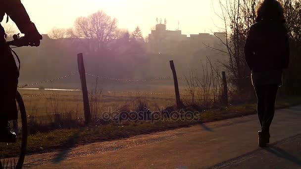 Een Man die rijden op een fiets en een jong meisje, wandelen langs aantal dorp weg in de zonnige stralen van een Sunrising in de herfst — Stockvideo