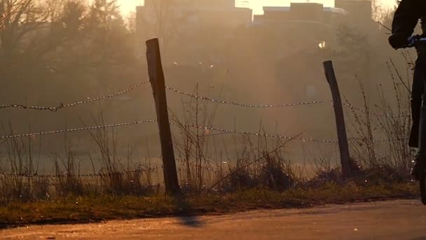 Een fietser met een aangezet koplicht rijdt langs een schilderachtige dorp weg in de stralen van de ochtend zon in Slow Motion. — Stockvideo