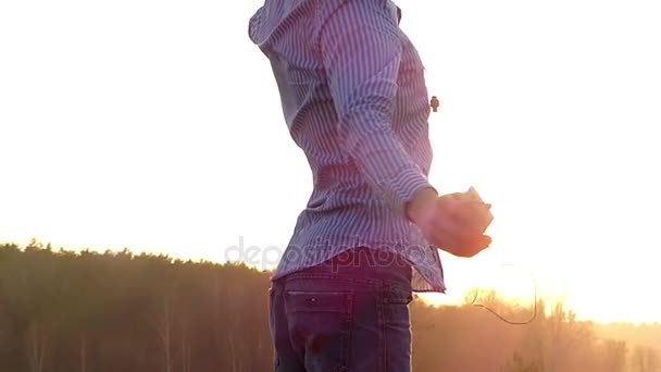 Jeune homme lève les mains et danse avec des écouteurs dans les oreilles et un joueur dans les mains avec les rayons ensoleillés rayonnant de derrière à Slo-Mo . — Video