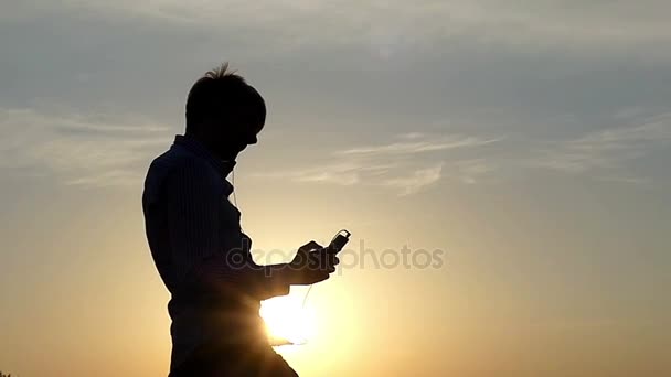 Happy Young Man está dançando com fones de ouvido em suas orelhas e um jogador em sua mão com o céu cinzento nublado no fundo no início da primavera — Vídeo de Stock