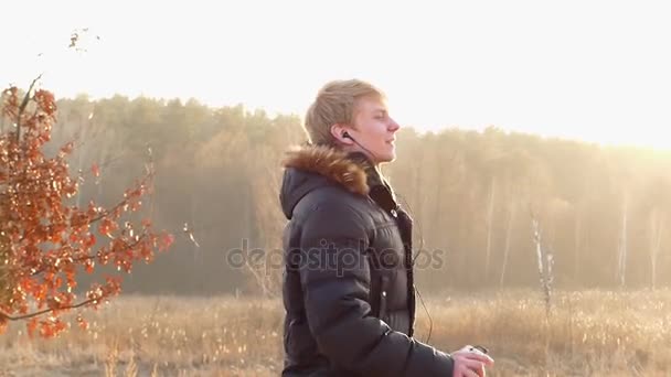 Молодой человек танцует в стиле диско с наушниками в ушах на впечатляющем закате на открытом воздухе поздней осенью в медленном движении — стоковое видео
