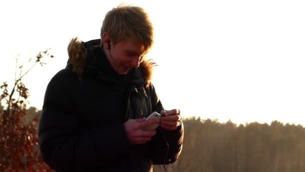 Hombre joven está mirando un teléfono inteligente con auriculares en sus oídos en el impresionante atardecer al aire libre en una primavera temprana — Vídeo de stock