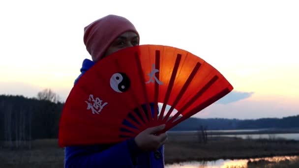 Mulher bonita está acenando com seu ventilador portátil para trás e para a frente ao ar livre no banco de um lago no início da primavera — Vídeo de Stock