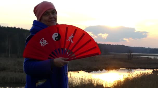 Bella donna sta agitando avanti e indietro con un orientale arancione palmare ventilatore nei raggi del tramonto e sorrisi sulla riva di un lago foresta — Video Stock
