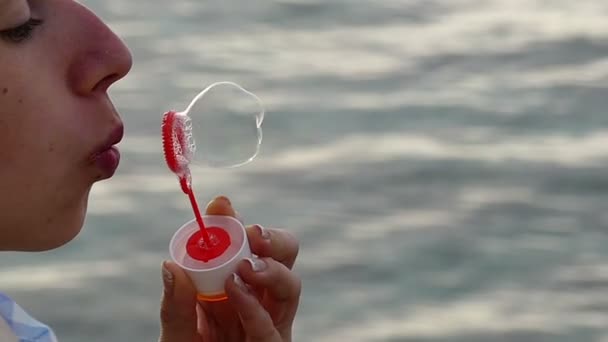 Joven hermosa chica está creando burbujas de jabón con una varita de jabón rojo de una manera divertida con agua azul del mar en el fondo — Vídeo de stock