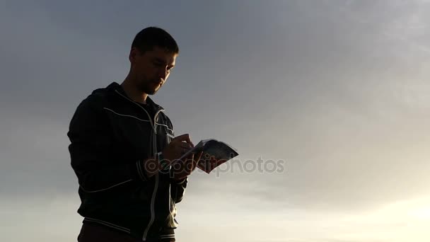 En silhuett av en människa skriver ner något i sin anteckningsbok och fin ser solnedgången. — Stockvideo