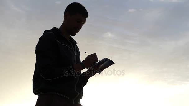 Man schrijven iets in zijn notitieblok bij zonsondergang. — Stockvideo